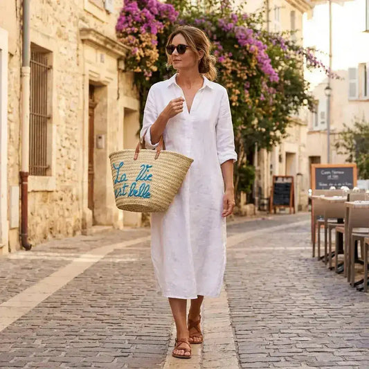 Woman in white linen dress walking on cobblestone street with straw tote bag and sunglasses