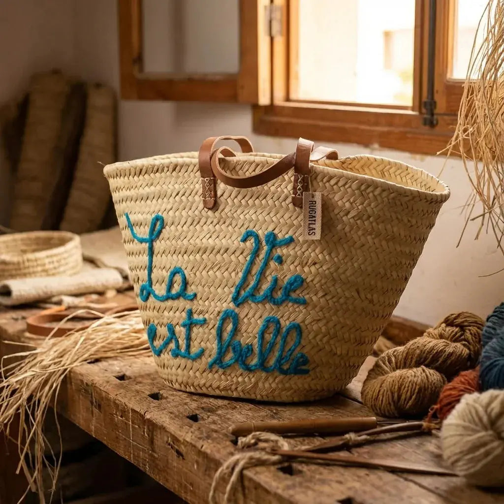 Handwoven straw basket with leather handles and blue embroidered French phrase on wooden table