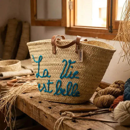 Handwoven straw basket with leather handles and blue embroidered French phrase on wooden table