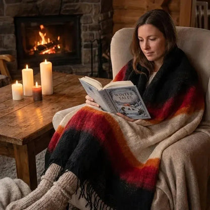 Femme lisant 'Winter Tales' dans un fauteuil près d'une cheminée avec plaid coloré et bougies