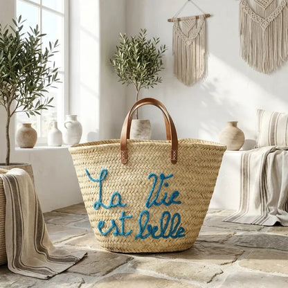 Woven straw tote bag with leather handles and blue embroidery of La Vie est Belle in sunlit boho room