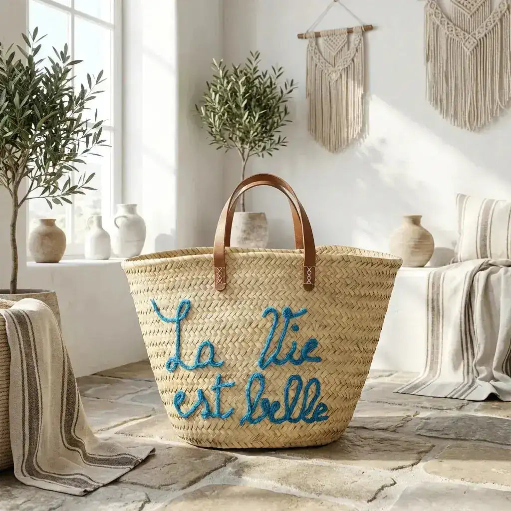 Woven straw tote bag with leather handles and blue embroidery of La Vie est Belle in sunlit boho room