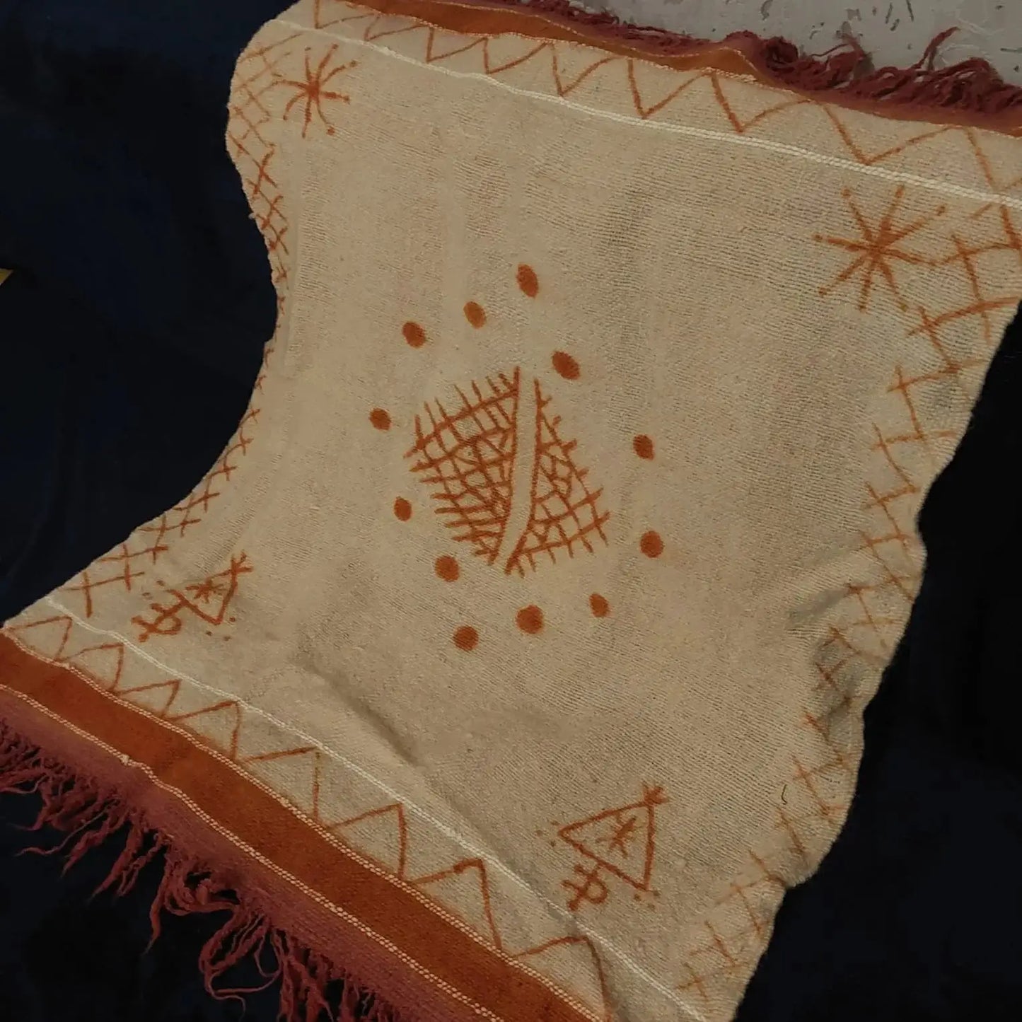 Beige woven textile with rust orange geometric and star patterns, and fringed edges on dark fabric
