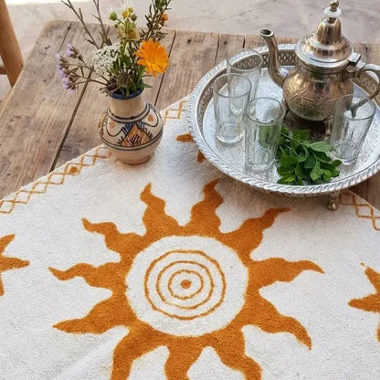 Tapis blanc avec motif soleil orange, plateau en métal avec théière, verres et menthe, vase avec fleurs sur table en bois