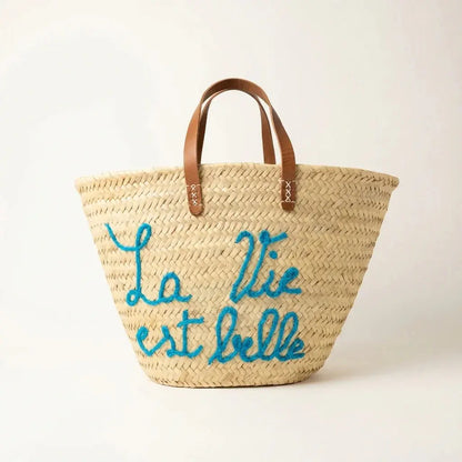 Woven straw tote bag with brown leather handles and blue embroidered French phrase La Vie est belle