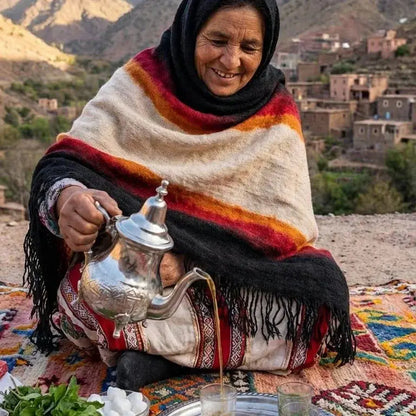 Femme berbère en tenue traditionnelle versant du thé d'une théière en argent, décor montagne et village en arrière-plan