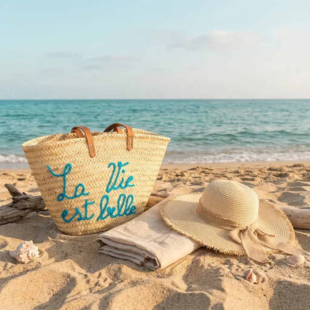 Straw beach bag with blue text next to sun hat and towel on sandy shore by calm sea