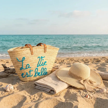Straw beach bag with blue text next to sun hat and towel on sandy shore by calm sea