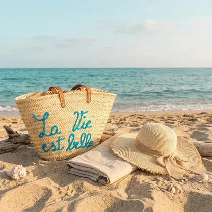 Straw beach bag with blue text next to sun hat and towel on sandy shore by calm sea