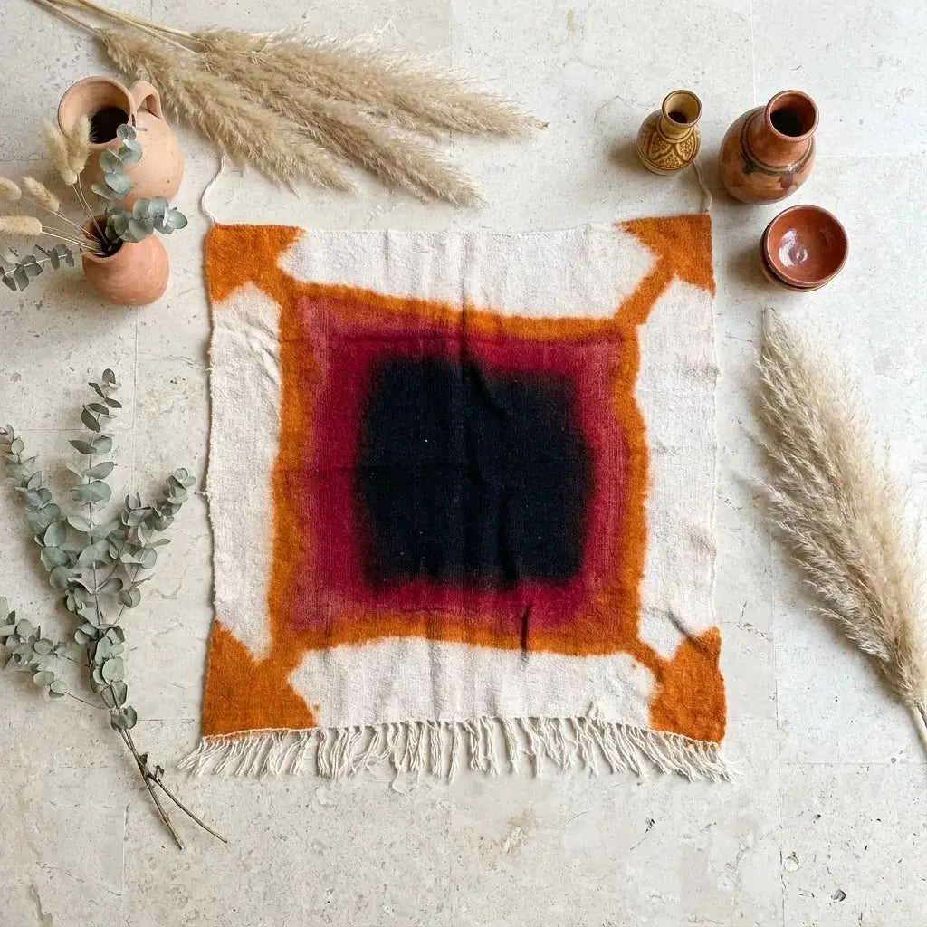 Tapis décoratif carré en laine avec motifs géométriques orange, rouge et noir, décor bohème avec vases en terre cuite et plantes sèches