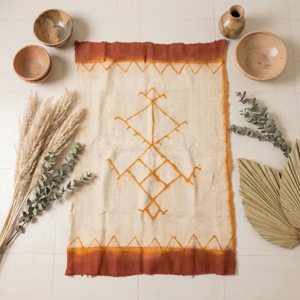 Handwoven Moroccan rug with orange geometric design, surrounded by pottery, pampas grass, eucalyptus, and dried palm leaves