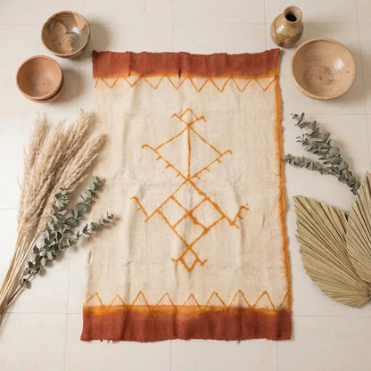 Handwoven Moroccan rug with orange geometric design, surrounded by pottery, pampas grass, eucalyptus, and dried palm leaves