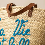 Close-up of woven straw tote bag with leather handles and blue embroidered text