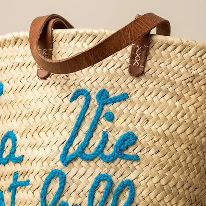 Close-up of woven straw tote bag with leather handles and blue embroidered text